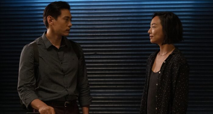 Escena nocturna de Past Lives (2023, A24) donde Hae Sung (Teo Yoo) y Nora (Greta Lee) mantienen una conversación íntima. Los protagonistas se miran de perfil con intensidad, él viste una camisa a cuadros y ella un conjunto negro con detalles brillantes, mientras un fondo de persianas azules metálicas crea una atmósfera urbana y melancólica característica de la película.