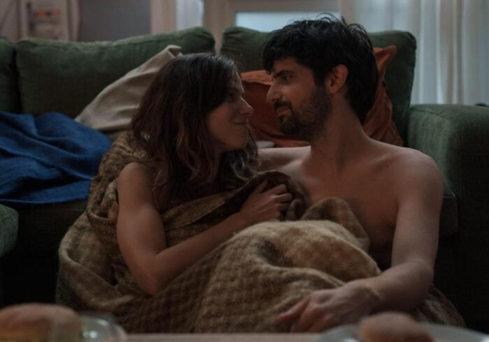 Escena íntima de Los Años Nuevos (Movistar Plus+) donde Ana (Iria del Río) y Óscar (Francesco Carril) comparten un momento de complicidad en el sofá. La pareja protagonista aparece recostada bajo una manta marrón, con un sofá verde de fondo y una iluminación tenue que enfatiza la intimidad del momento. Sus miradas cercanas y gestos naturales capturan la cotidianidad del amor que retrata la serie