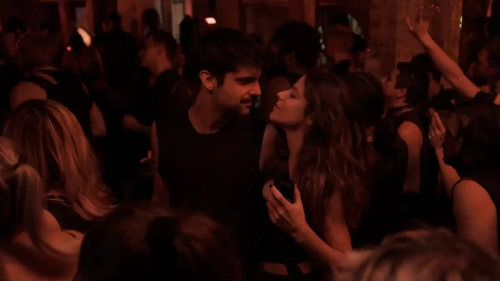 Escena íntima de Los Años Nuevos (Movistar Plus+) donde Ana (Iria del Río) y Óscar (Francesco Carril) comparten una mirada cómplice en medio de una discoteca llena de gente bailando. La imagen captura la intimidad de su relación incluso en un espacio público y ruidoso, mostrando cómo su conexión trasciende el entorno social. El contraste entre la energía del ambiente festivo y la quietud de su momento personal define la esencia de la serie.