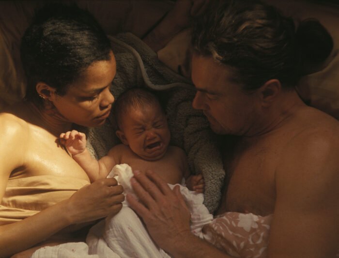 Willa, una recién nacida, en brazos de Ghetto y Perfidia Beverly Hills mientras descansan juntos en una cama, en una escena íntima y cálida