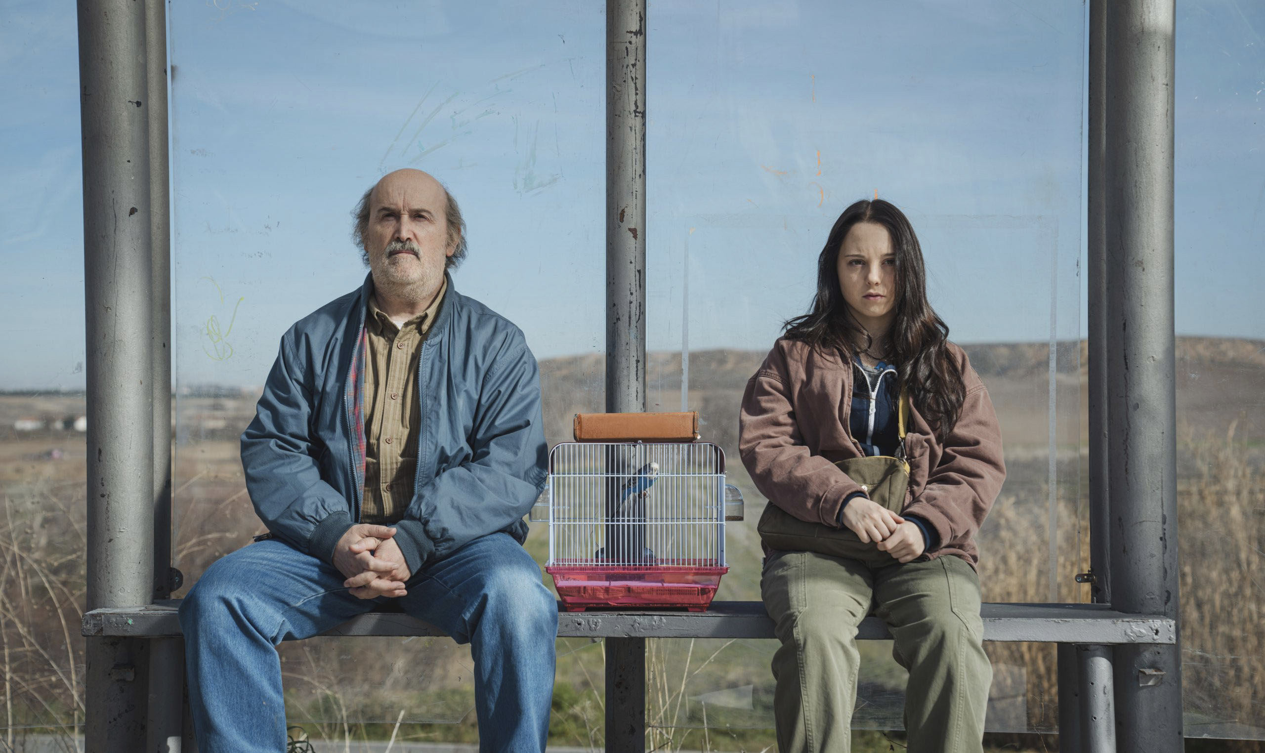 Joserra y Mar, personajes de Yakarta de Movistar Plus+, interpretados por Javier Cámara y Carla Quílez, sentados en una parada de autobús junto a una jaula con un pájaro